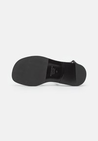 Musta korkokenkä, jonka pohja on sileä, hieman pyöristetty muotoilu ja minimalistinen design. Painettu brändi lähellä kantapäätä.