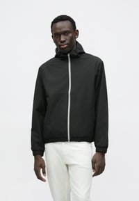 Μαύρο αδιάβροχο μπουφάν με κουκούλα, εμπρόσθιο φερμουάρ και ελαστικές μανσέτες. Διαθέτει αντίθετες λευκές κάθετες ρίγες κατά μήκος του φερμουάρ.