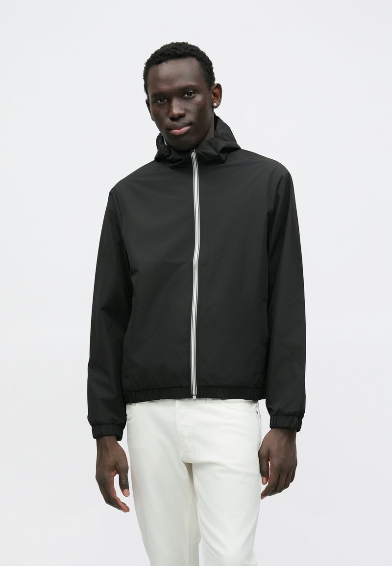 Μαύρο αδιάβροχο μπουφάν με κουκούλα, εμπρόσθιο φερμουάρ και ελαστικές μανσέτες. Διαθέτει αντίθετες λευκές κάθετες ρίγες κατά μήκος του φερμουάρ.