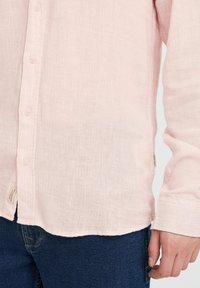 Chemise boutonnée rose clair en tissu texturé avec un motif côtelé. Présente un ourlet arrondi et est assortie à un jean bleu foncé.