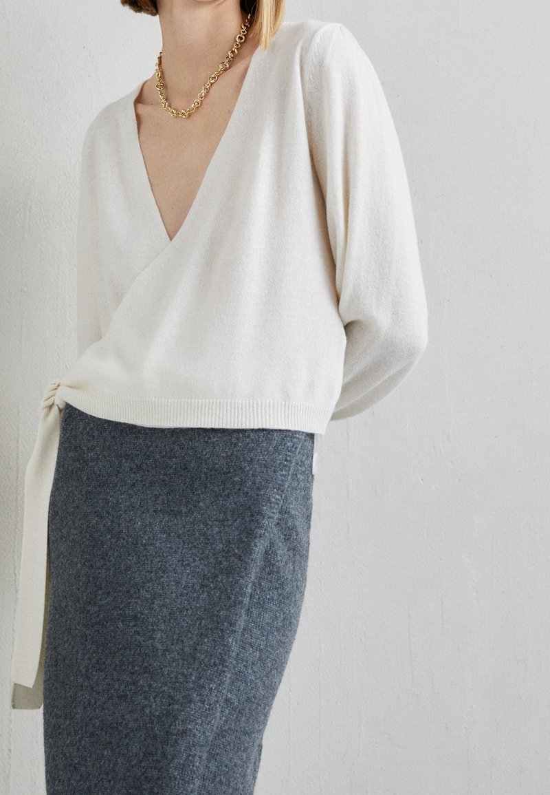 Femme portant un pull cache-cœur blanc à col en V noué sur le côté, avec une jupe moulante gris foncé et un collier chaîne doré.
