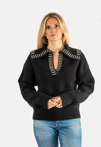 Maglione nero a maglia con scollatura a V profonda, colletto largo con borchie e polsini a coste. Abbinato a jeans azzurri chiari, con le mani incrociate davanti.