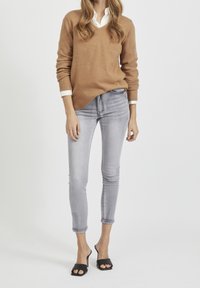 Lichtbruine gebreide trui met een V-hals, gecombineerd met een paar grijze skinny jeans en zwarte hakken. Platte textuur, casual pasvorm en strakke lijnen.