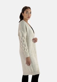 Lång, krämfärgad stickad cardigan med ribbad textur, kabelstickade ärmar och öppen frontdesign. Bärs över en svart topp.