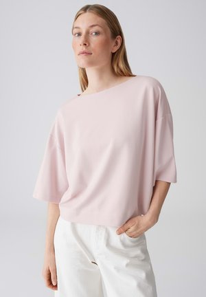 Donna con capelli biondi lisci indossa una maglietta rosa pallido a maniche corte e pantaloni bianchi, in piedi con una mano in tasca contro uno sfondo uniforme.