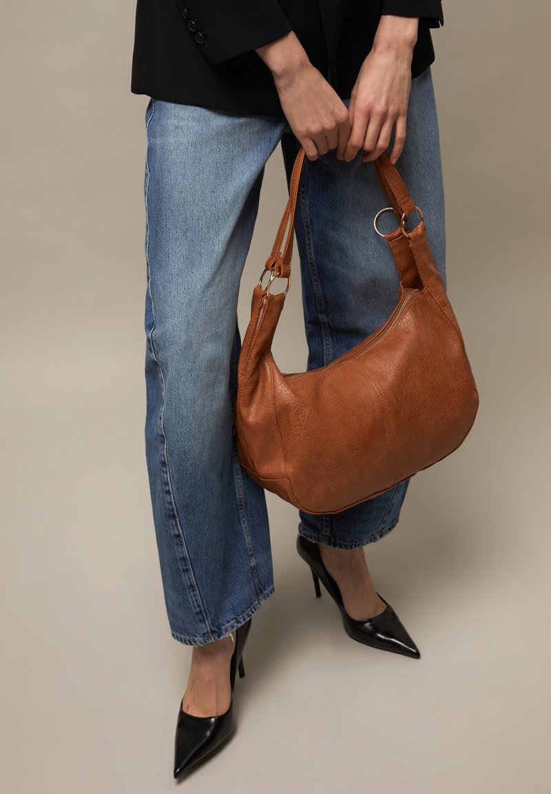 Persona che indossa jeans blu e tacchi alti neri che tiene una borsa in pelle marrone con doppi manici e anelli metallici.