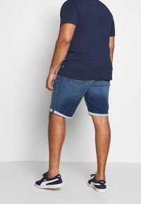 Muž v námořnickém modrém tričku, s vyhrnutými denimovými kraťasy a námořnickými teniskami Puma stojí zády ke kameře u bílé zdi.