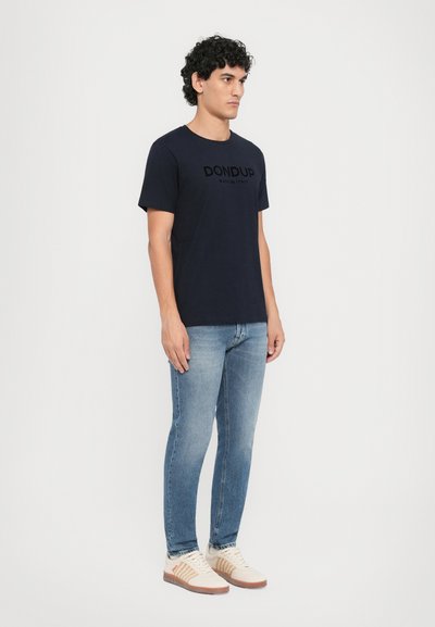 Navy blue cotton t-shirt with short sleeves displaying a black printed logo, paired with light blue slim-fit denim jeans and beige sneakers.