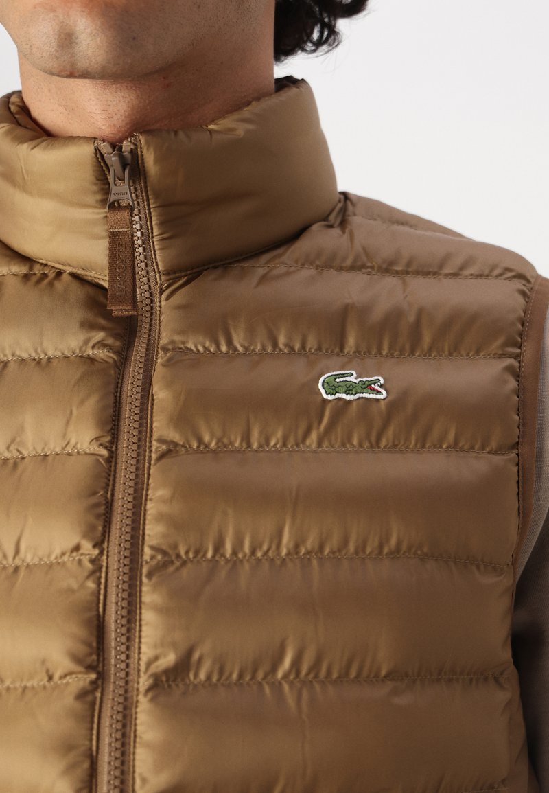 Braune Steppweste mit horizontaler Steppung, hohem Kragen und Reißverschluss vorne. Mit einem kleinen grünen Krokodil-Logo auf der Brust.