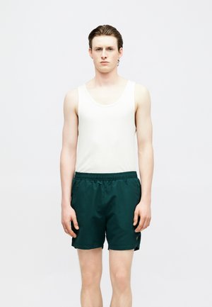 Νεαρός άνδρας που στέκεται με το πρόσωπο προς τα εμπρός, φορώντας λευκή αμάνικη μπλούζα και σκούρα πράσινα σορτς με ελαστική μέση, μπροστά σε απλό φόντο.