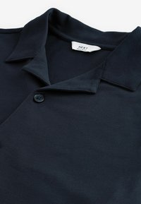 Polo de color marino hecho de una tela texturizada, con un collar clásico, un solo botón negro y una etiqueta de marca cosida dentro del collar.