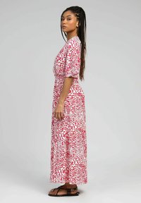Robe maxi blanche avec un motif éclaboussé rouge et rose, manches courtes bouffantes, taille cintrée et jupe évasée. Portée avec des sandales marron.