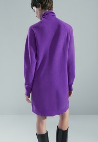 Personne portant une longue robe-pull à col roulé violette et des bottes noires montantes jusqu'aux genoux, debout devant un fond gris uni.