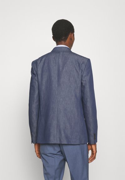 Férfi, aki illeszkedő kék zakót és hozzá illő nadrágot visel, háttal áll a néző felé egy sima, világos háttér előtt.