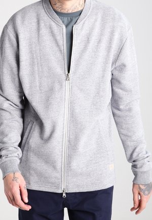 Homme portant une veste zippée gris clair sur une chemise bleue et un pantalon foncé, montrant des tatouages sur le cou et les mains, debout devant un fond uni.