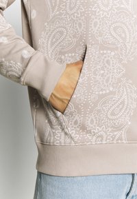 Felpa leggera beige chiaro con motivo paisley bianco, polsini a coste e tasca a marsupio. Presenta un tessuto testurizzato e una vestibilità comoda.