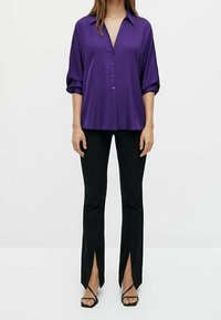 Lilla satin langærmet skjorte med knapper foran, kombineret med sorte bukser med slidser i benene. Model iført sorte sandaler med stropper.