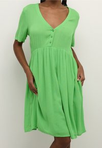 Grünes Kleid mit kurzen Ärmeln, V-Ausschnitt, Knopfdetail an der Brust und einem gerafften Rock. Der Stoff hat ein weiches, strukturiertes Aussehen.