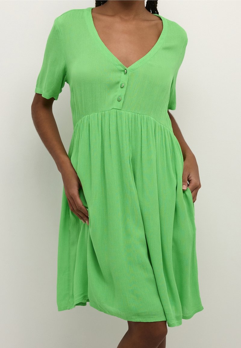 Grünes Kleid mit kurzen Ärmeln, V-Ausschnitt, Knopfdetail an der Brust und einem gerafften Rock. Der Stoff hat ein weiches, strukturiertes Aussehen.