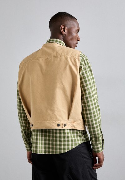 Tan cotton vest with a cropped design. The back features a smooth texture and two snap buttons at the hem. Below, a green and white checkered shirt.