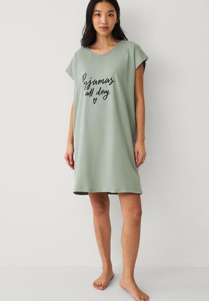 Vrouw met lang donker haar die een loszittende groene nachtjapon draagt met de tekst "Pyjama's de hele dag" en een klein hartje, staand op blote voeten tegen een effen achtergrond.