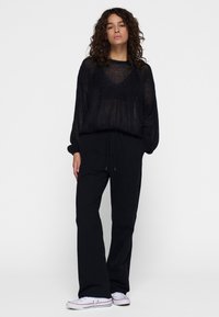 Zwarte doorzichtige lange mouw top met een V-hals en een ontspannen pasvorm, gecombineerd met zwarte broek met trekkoord en witte sneakers met kleuraccenten.
