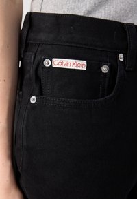 Tasca dei jeans in denim nero con etichetta bianca e rossa di Calvin Klein, rivetti argentati e una maglia a costine grigia chiara infilata dentro.