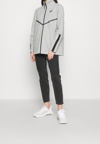 Giacca grigia con zip, dettagli neri e colletto alto, abbinata a jeans neri slim-fit e sneakers bianche con dettagli rosa.