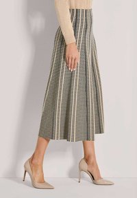 Frau in beiger Langarmbluse und hoch tailliertem, gemustertem Midikleid mit beigen, spitzen High Heels, die seitlich steht.