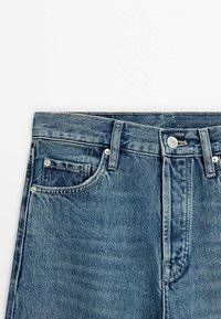 Blå denimjeans med hög midja, fem fickor, metallnitar och knappstängning. Har en blekt, texturerad yta.