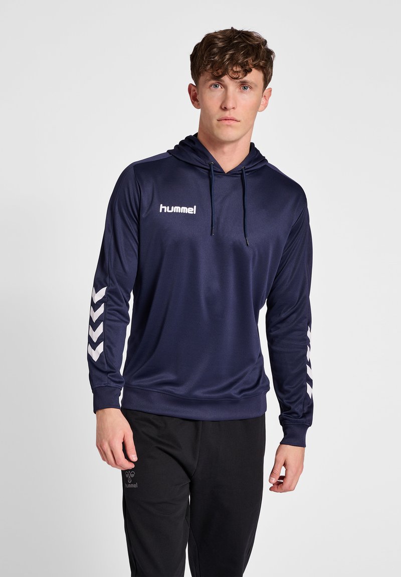 Jonge man draagt een donkerblauwe hummel hoodie met chevronstrepen op de mouwen en zwarte hummel broek, staand tegen een effen lichte achtergrond.