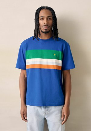 Mann mit schulterlangen Dreadlocks, der ein blaues T-Shirt mit grünen, weißen und orangenen horizontalen Streifen trägt, steht vor einem beige-farbenen Hintergrund.