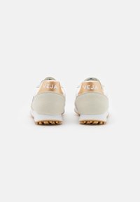 Hvite sneakers med en teksturert, lys grå suedeflapp på hælen. Har et brunt logo-accent. Gummisåle med små gripeknubber.