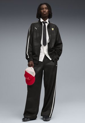 Nuori henkilö punotut hiukset, musta Puma-verryttelypuku, valkoinen paita, musta solmio, pitämässä puna-valkoista lakkia, seisoo harmaata taustaa vasten.