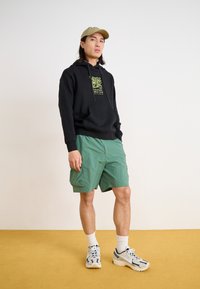 Black hoodie with a graphic design, green shorts, and white sneakers with blue accents. Model wears a beige cap and white socks.
