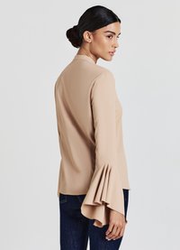 Blusa beige con colletto alto e maniche svasate. Tessuto morbido, silhouette aderente e design minimalista, abbinata a jeans blu scuro.