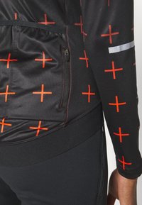 Sort sporttop med røde plus-tegn, med en lynlåsplads og reflekterende stribe. Lavet af glat, strækbart stof.