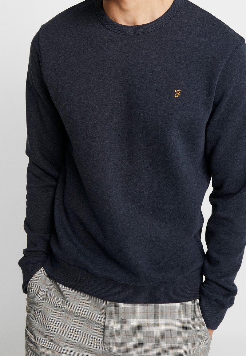 Homme portant un pull bleu marine foncé à col rond avec un petit logo jaune "F" sur la poitrine et un pantalon à carreaux gris, les mains dans les poches.