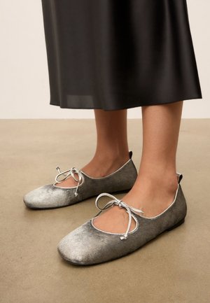 Persoon draagt zilverkleurige fluwelen ballerina's met strikjes, staand op een beige vloer, met een zwarte rok zichtbaar boven de enkels.