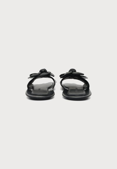 Paire de sandales plates noires avec des brides nouées, vues de dos sur un fond gris clair uni.