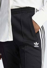 Černé sportovní kalhoty s bílými postranními pruhy, vyrobené z hladkého materiálu. Mají elastický pas a logo Adidas na levém boku.