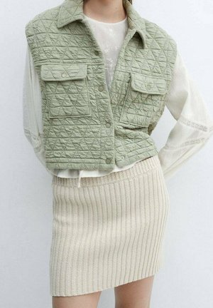 Veste matelassée sans manches vert sauge avec fermetures à pression, portée sur un haut blanc à manches longues et une mini-jupe crème en maille côtelée.