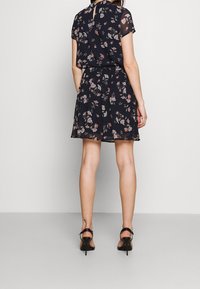 Vestido floral navy con mangas cortas, que presenta un lazo en el cuello y una cintura ajustada, falda fluida y textura transparente. Tacones altos negros.