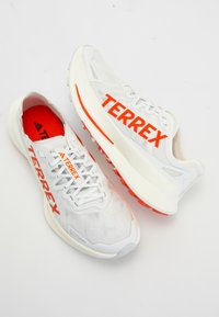 Białe buty biegowe z pomarańczowym logo "TERREX", z górą z siateczki, amortyzowaną podeszwą i teksturowanym bieżnikiem z gumy dla lepszej przyczepności. Lekka konstrukcja.