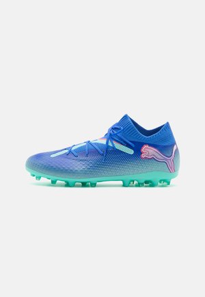 Botín de fútbol sintético azul con upper texturizado, diseño ligero, suela verde y logo de Puma rosa en el lateral.