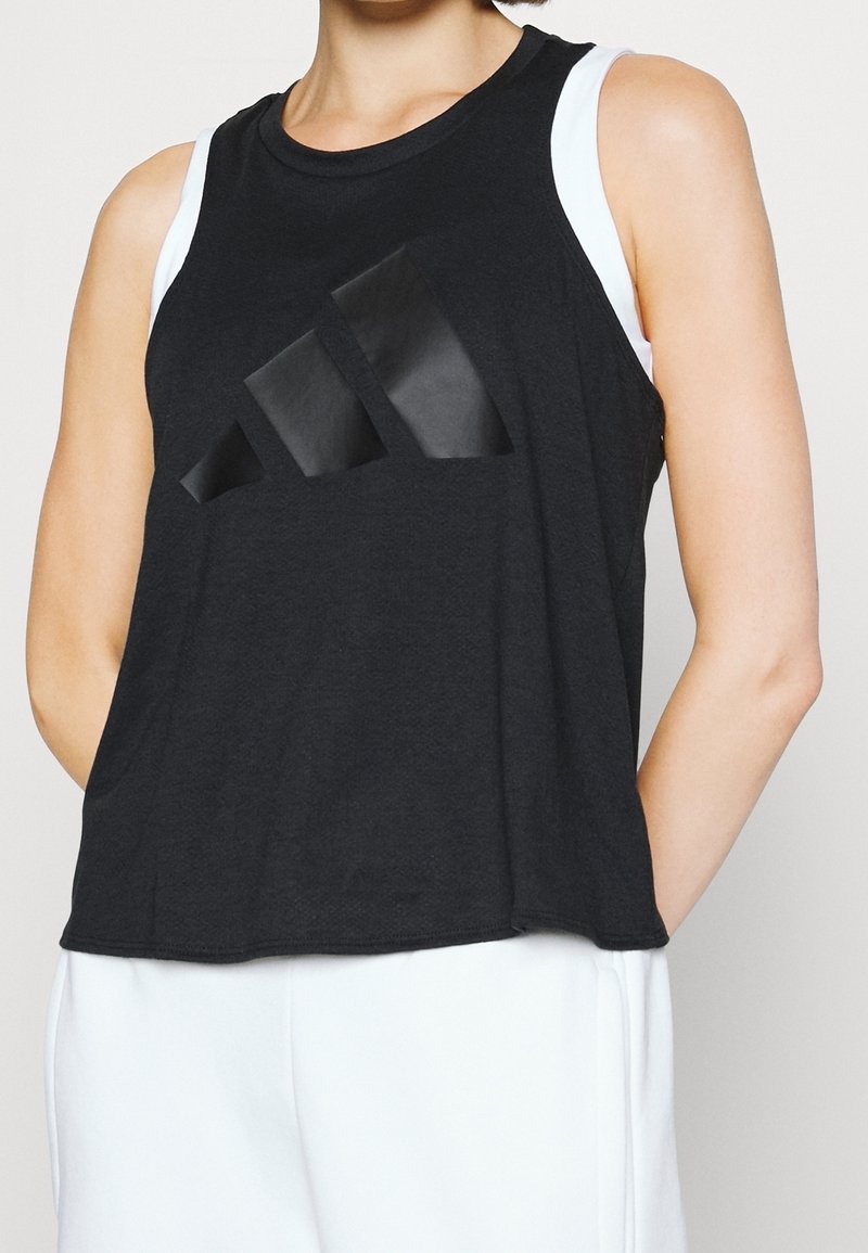 Femme portant un haut noir sans manches Adidas avec un grand logo noir et un pantalon blanc, debout devant un fond uni.