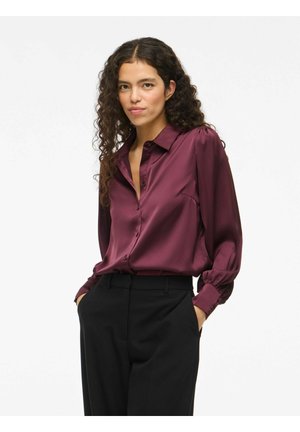 Chemisier en satin bordeaux avec un col, un devant boutonné et des manches longues regroupées au niveau des poignets. Assorti à un pantalon noir cintré.