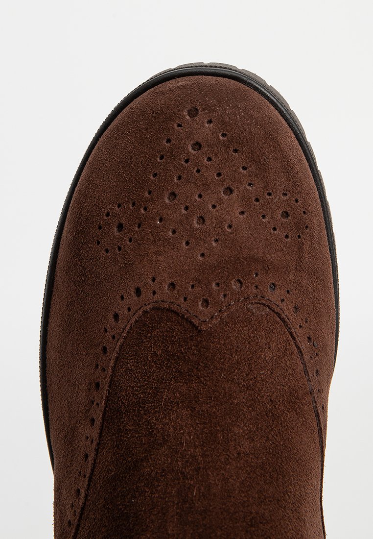 Bota de ante marrón con detalles perforados, bordes festoneados y suela negra plana. Presenta una textura suave y un diseño de punta redondeada.
