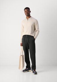 Haut à manches longues beige clair à col zippé, pantalon gris foncé avec cordon de serrage, baskets noires et bleues, portant un tote bag beige.