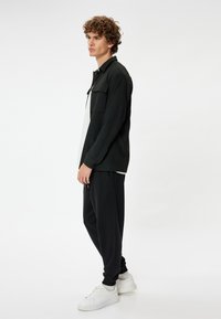 Sort strikket jakke med krave og forlommer, båret over en hvid skjorte; parret med sorte sweatpants og hvide sneakers.